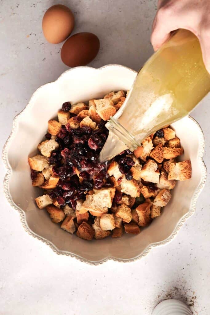 Croutons and cranberries sit in a bowl as a hand pours liquid from a bottle, preparing the perfect bread stuffing. Two eggs rest nearby on the light surface, ready to join the mix.
