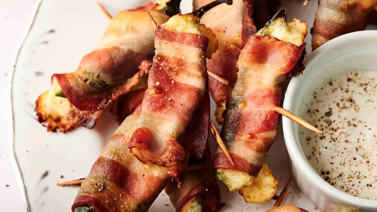Bacon-wrapped jalapeños filled with cheese, secured with toothpicks, on a plate next to a small bowl of dipping sauce.