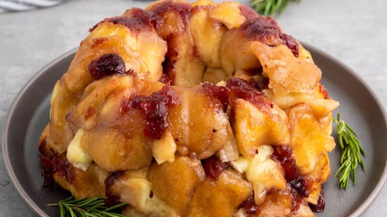 A baked pull-apart bread topped with melted cheese and cranberry sauce, garnished with rosemary sprigs on a plate.