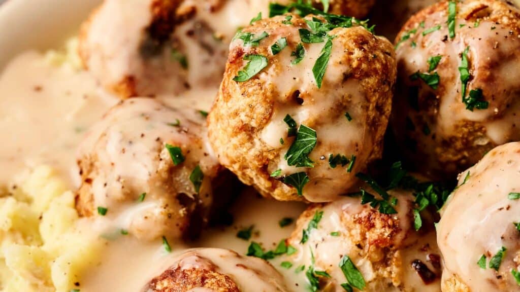 Plate of meatballs with creamy sauce and chopped herbs on top, served over mashed potatoes.