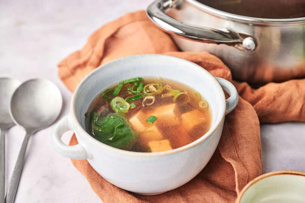 A steaming bowl of miso soup, rich with tofu, seaweed, and green onions, sits invitingly on a cloth alongside a gleaming stainless steel pot and two spoons.