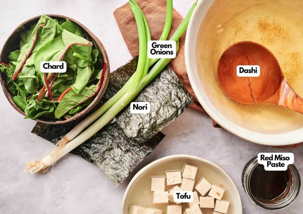 Bowls and plates contain chard, green onions, nori seaweed, tofu cubes, red miso paste, and a bowl of dashi with a ladle—all the fresh ingredients you need to prepare classic miso soup.