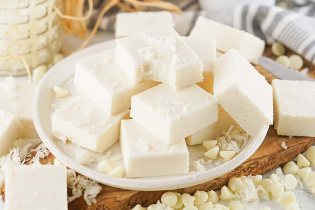 Creamy white coconut fudge squares piled on a white plate on a wooden board, garnished with shredded coconut and surrounded by white chocolate chips