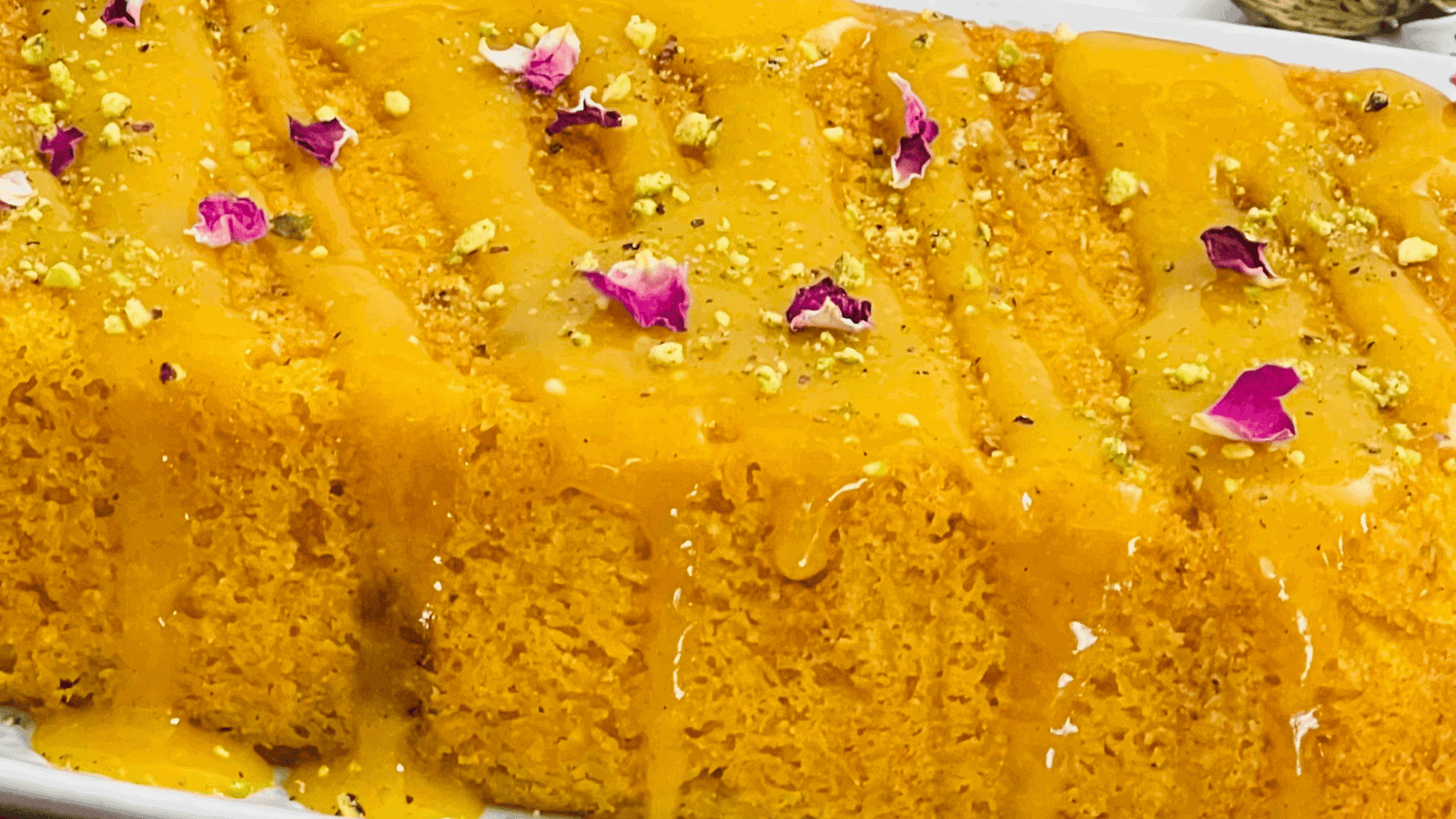 Close-up of a vibrant orange dessert garnished with flower petals and nuts, drizzled with syrup on a white rectangular plate.