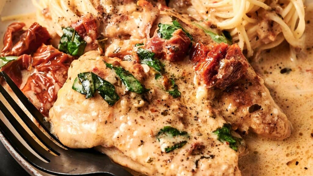 Creamy chicken dish with sun-dried tomatoes, spinach, and black pepper, served with spaghetti on a plate, with a fork on the side.