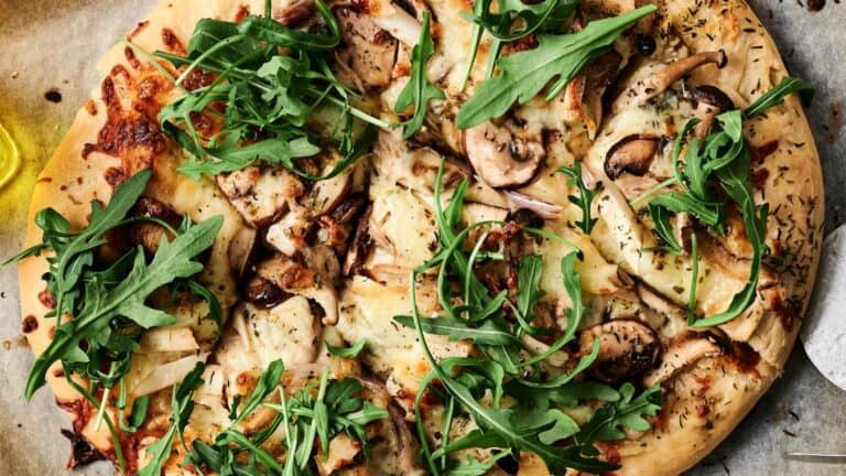 A pizza topped with mushrooms, arugula, cheese, and herbs on a wooden surface.