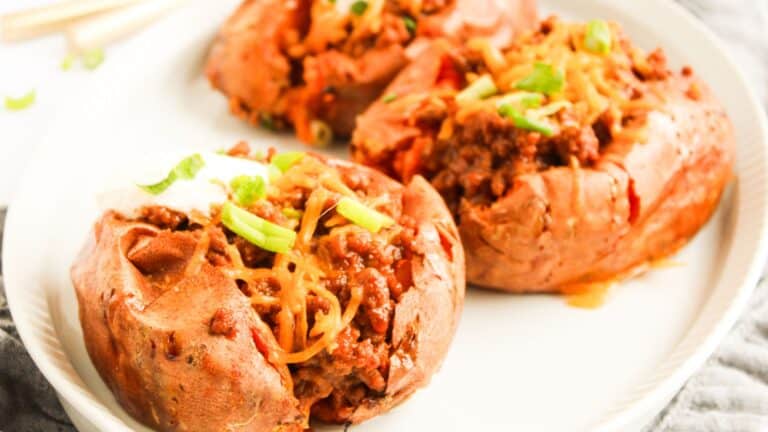 Three baked potatoes topped with chili, melted cheese, sour cream, and chopped green onions on a white plate.