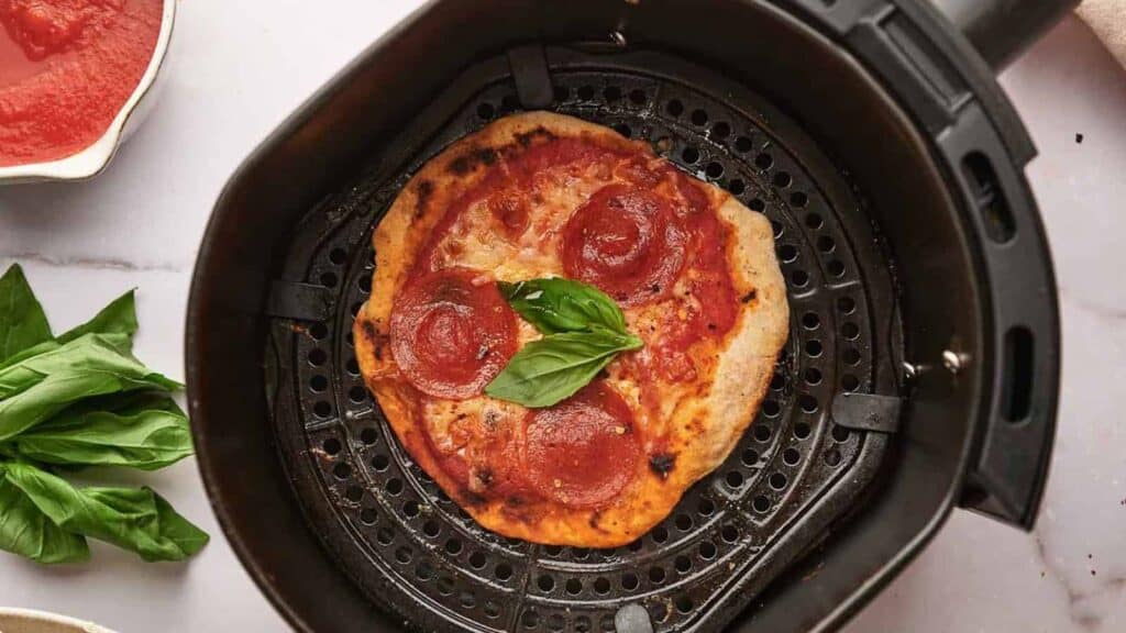A pepperoni pizza with basil on top is inside an air fryer. Fresh basil leaves and a bowl of tomato sauce are nearby.