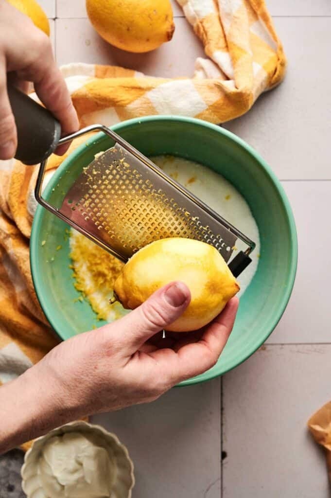 Hands grating lemon zest into a green bowl, with a yellow checkered cloth and another lemon nearby on a tiled surface, perfectly setting the stage for creating a delightful lemon pound cake.