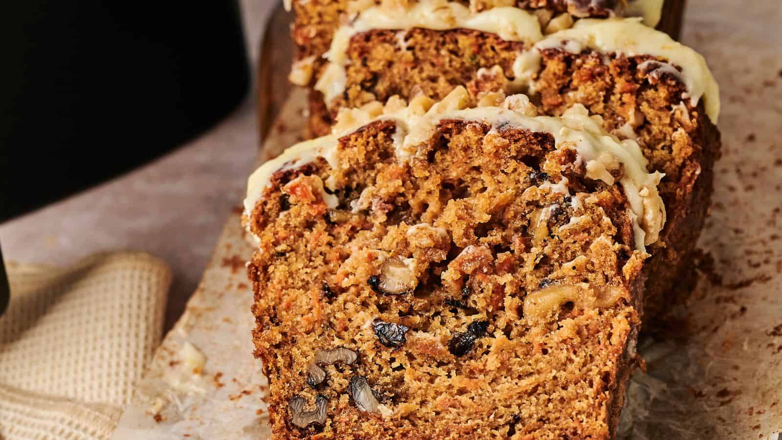 Sliced carrot cake with nuts is topped with cream cheese frosting, placed on a piece of parchment paper next to a napkin.