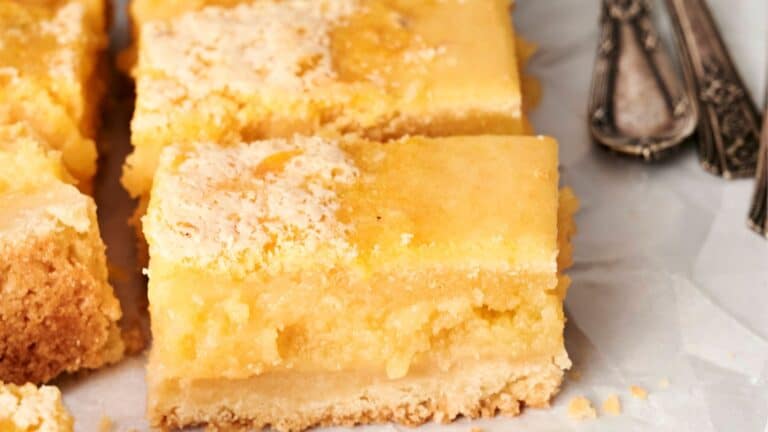Close-up of lemon bars with a crumbly base, a smooth lemon filling, and a light dusting of powdered sugar, arranged on parchment paper beside vintage metal cutlery.