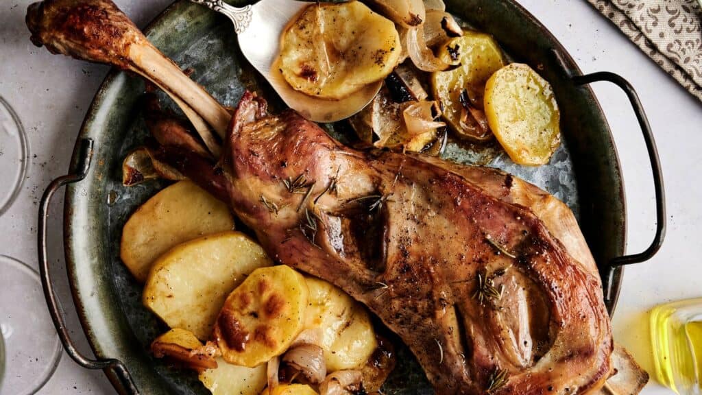 Roast lamb leg on a round metal tray, garnished with rosemary and served with sliced potatoes and onions.