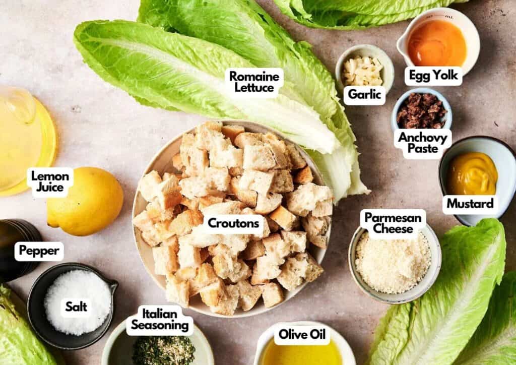 Caesar salad ingredients laid out on a light surface including romaine lettuce, croutons, egg yolk, garlic, anchovy paste, mustard, Parmesan cheese, lemon juice, olive oil, Italian seasoning, salt, and pepper