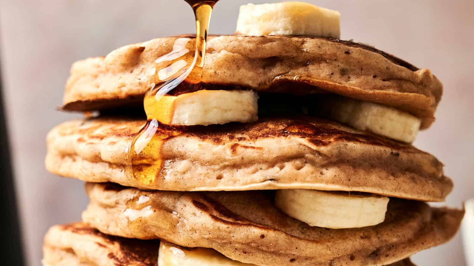 A stack of pancakes with banana slices between them, topped with syrup being poured from above.