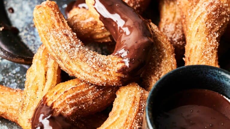 Close-up of churros covered in sugar and partially dipped in chocolate, arranged on a dark surface with a small bowl of chocolate sauce nearby.