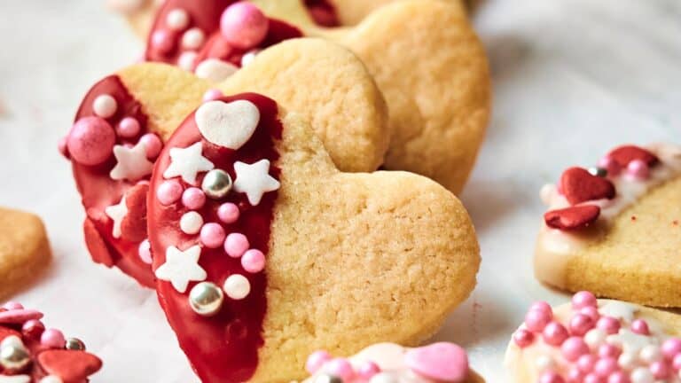 Heart-shaped cookies partially dipped in red icing, decorated with pink, white, and silver sprinkles and candy shapes.