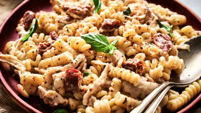 A plate of rotini pasta with shredded chicken, sun-dried tomatoes, creamy sauce, fresh basil, and a spoon on the side.