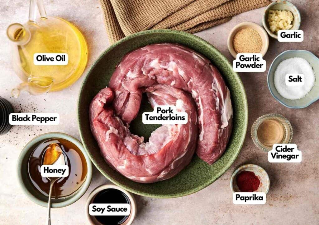 Pork tenderloin ingredients laid out on a light surface including raw pork tenderloins, olive oil, soy sauce, honey, garlic, garlic powder, paprika, cider vinegar, salt, and black pepper