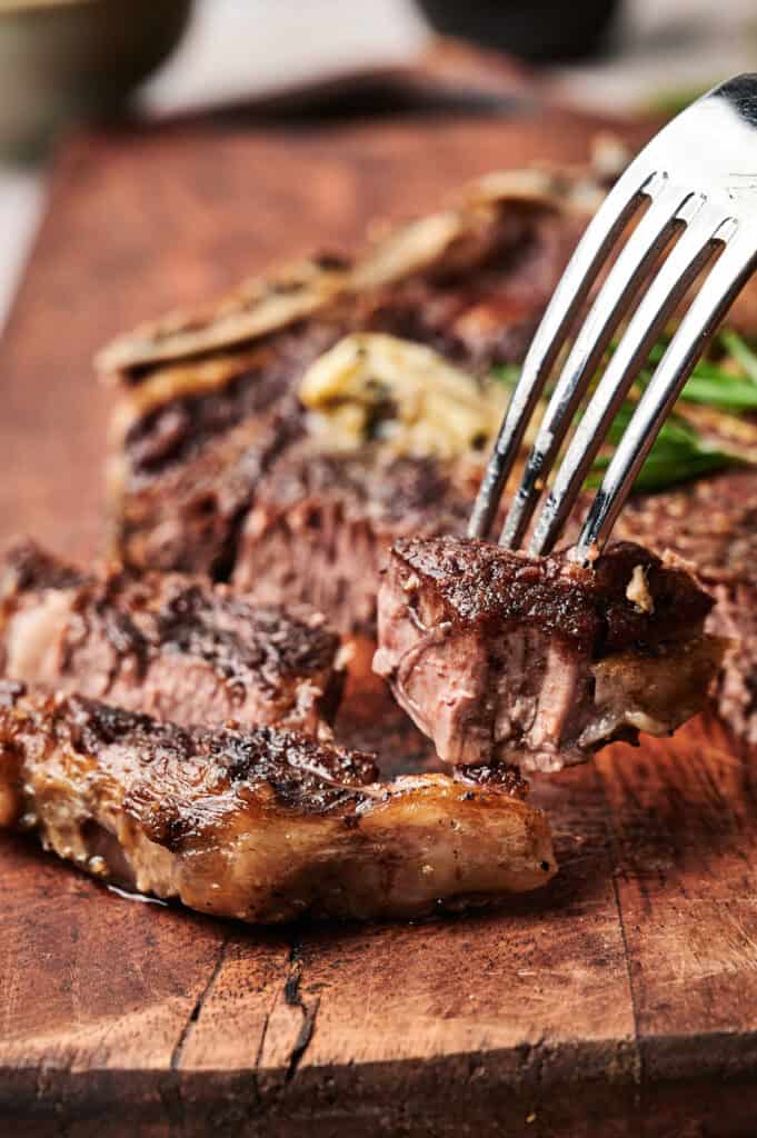 A fork holding a piece of grilled steak over a wooden cutting board with more steak pieces in the background.