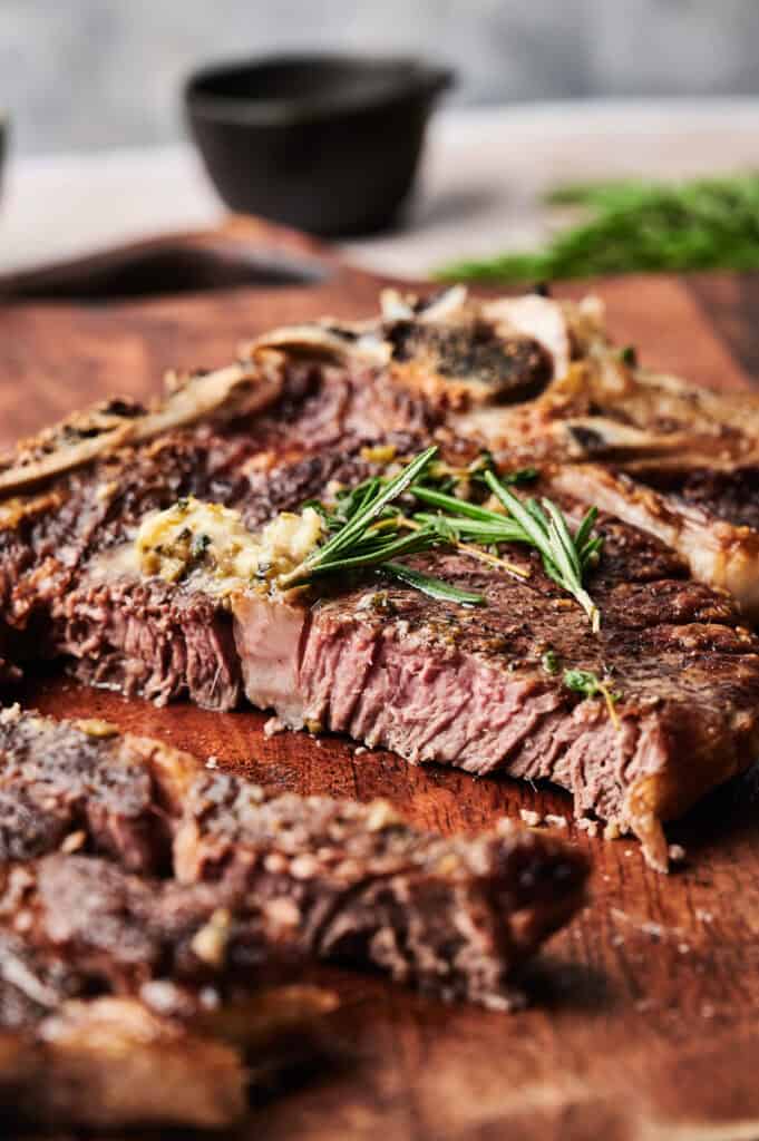 Grilled steak garnished with rosemary on a wooden cutting board, accompanied by a side dish in the background.