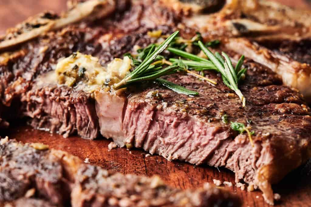 Close-up of a sliced, cooked steak topped with melted butter and fresh rosemary on a wooden surface.