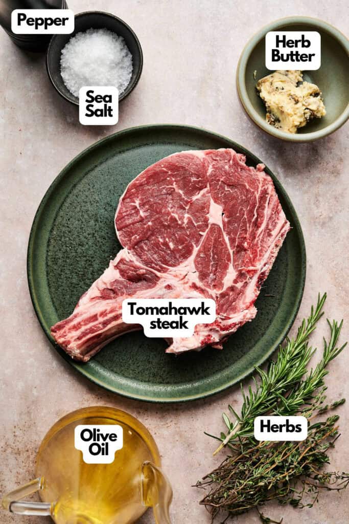 A raw tomahawk steak on a green plate, surrounded by herbs, olive oil, herb butter, sea salt, and pepper on a textured surface.