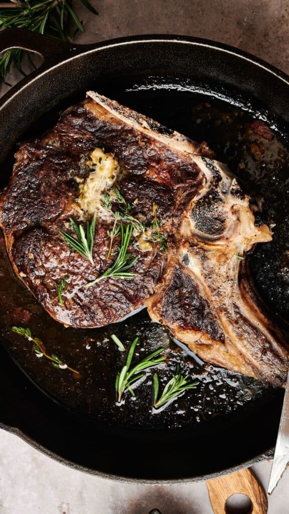 A cooked ribeye steak with herbs and butter in a cast iron skillet.