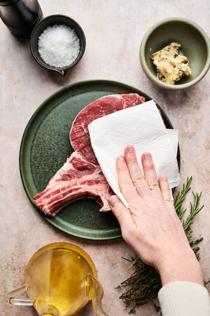 A hand pats a raw steak with a paper towel on a green plate. Nearby are bowls of salt, garlic butter, a rosemary sprig, and a glass container of oil.