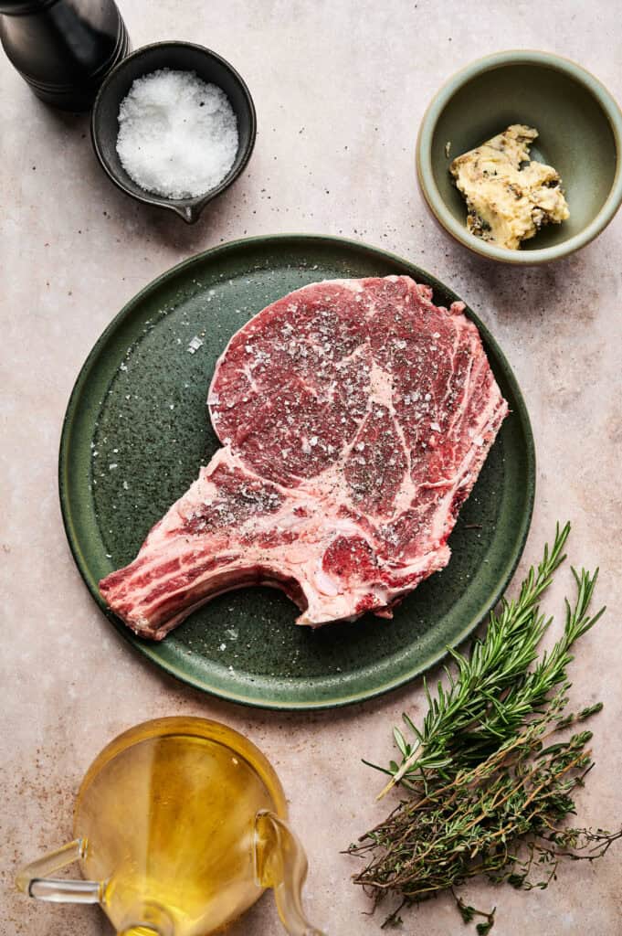 Raw tomahawk steak on a green plate, seasoned with salt, surrounded by rosemary, thyme, olive oil, minced garlic, and a bowl of salt on a beige surface.