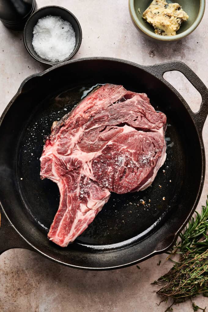 Raw, marbled steak sprinkled with salt and pepper in a cast iron pan. Nearby are bowls of salt and herb butter, with sprigs of fresh herbs on a light countertop.