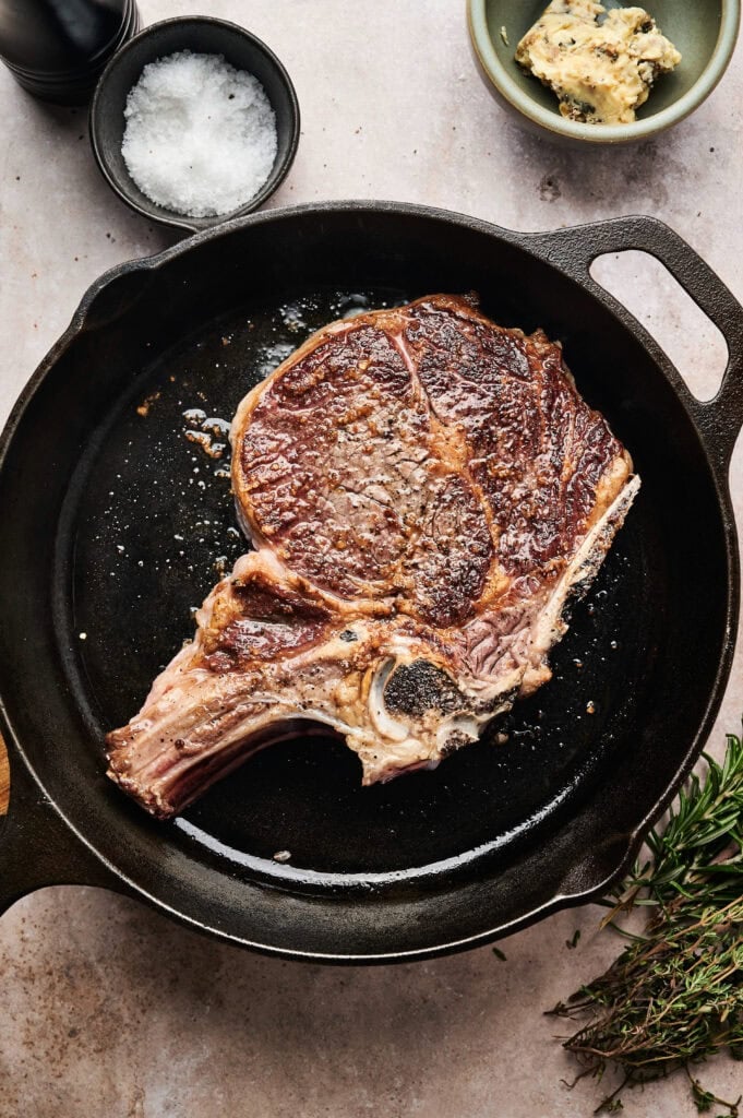 A cooked ribeye steak in a cast iron skillet, garnished with salt. A bowl of herb butter and salt are nearby, with a sprig of rosemary on the side.