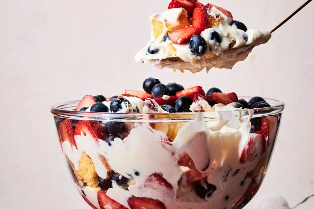 A large glass trifle bowl layered with pound cake, whipped cream cheese filling, fresh strawberries and blueberries, with a spoonful being lifted showing the red, white, and blue layers