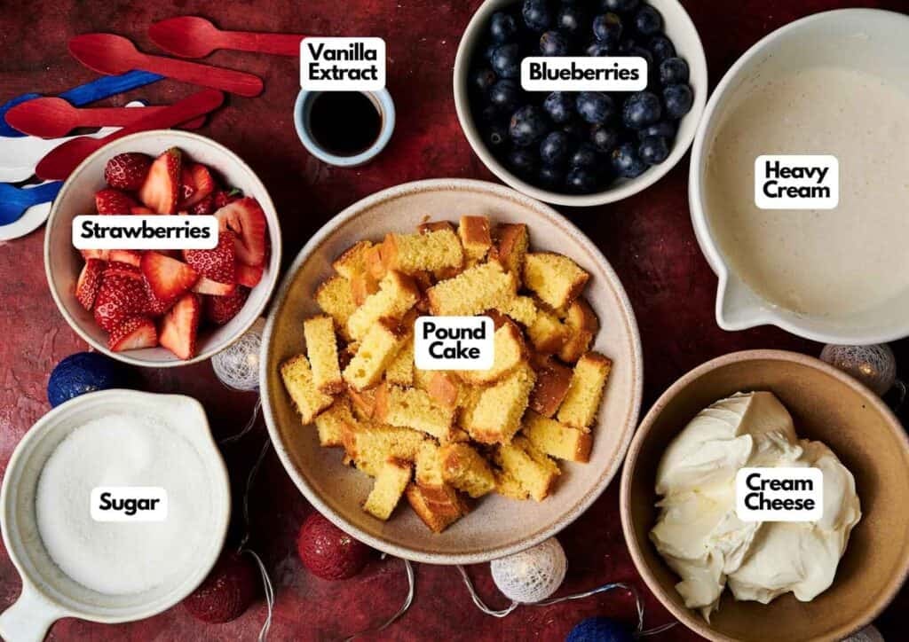 4th of July trifle ingredients laid out on a red surface including cubed pound cake, fresh strawberries, blueberries, cream cheese, heavy cream, sugar, and vanilla extract, with red, white, and blue decorations