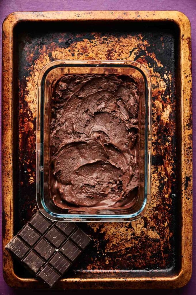 A glass dish filled with thick chocolate batter sits on a worn baking sheet, next to several squares of dark chocolate—perfect for making rich brownies or pairing with creamy chocolate ice cream.