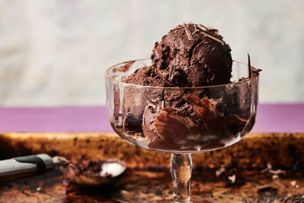 A glass dessert dish filled with two scoops of creamy chocolate ice cream, topped with chocolate shavings, sits on a rustic metal tray next to an ice cream scoop.