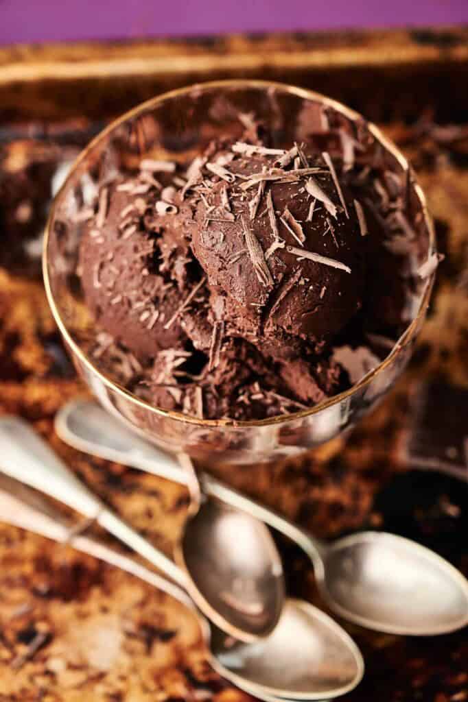 A glass bowl filled with scoops of rich chocolate ice cream, topped with chocolate shavings, sits next to three silver spoons on a rustic tray.