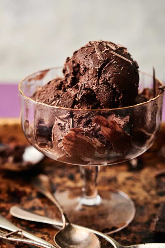 A glass dessert dish filled with scoops of rich chocolate ice cream, topped with delicate chocolate shavings, sits on a tray with spoons.