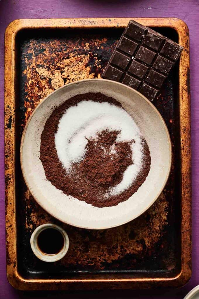 A bowl with cocoa powder and sugar, a bar of dark chocolate, and a small cup of vanilla extract on a worn baking sheet—classic ingredients for homemade chocolate ice cream.