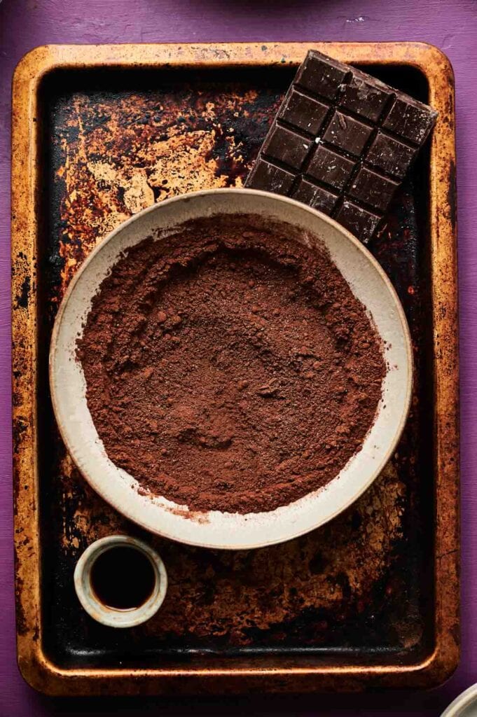 A bowl of cocoa powder, a bar of chocolate, and a small cup of liquid, perfect for making chocolate ice cream, are arranged on a worn baking sheet with a purple background.