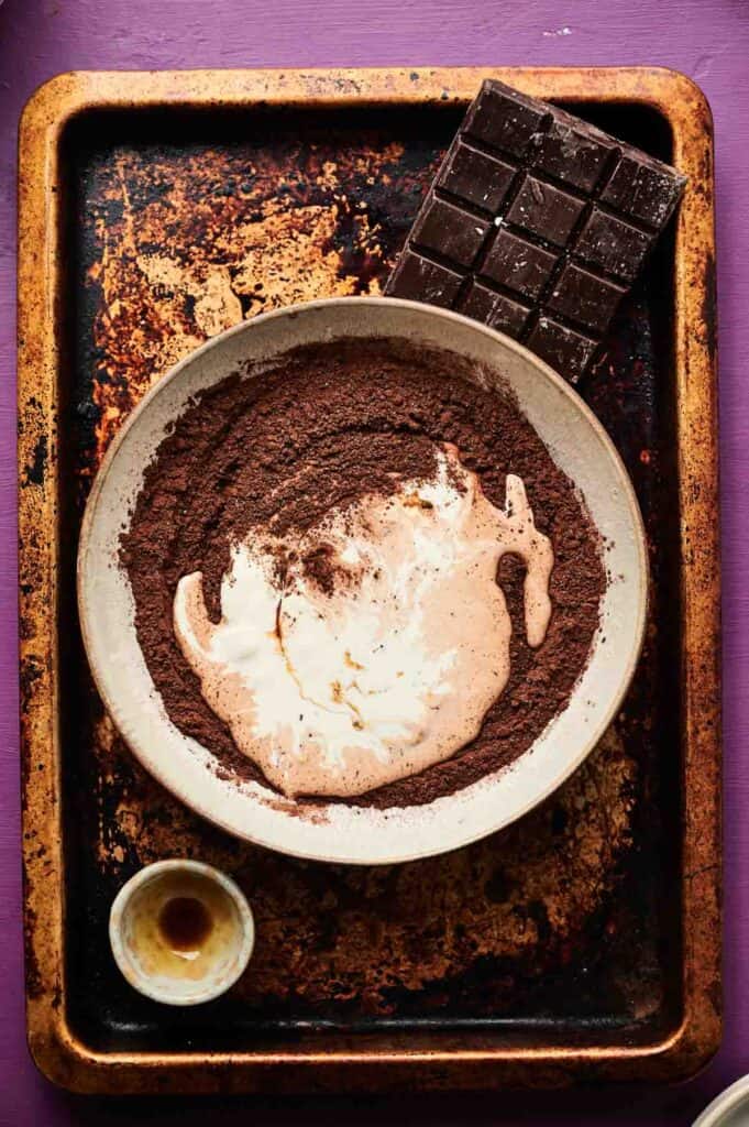A bowl with cocoa powder and cream sits on a worn baking tray next to a bar of dark chocolate and a small dish with liquid, ready for making rich chocolate ice cream, all set on a purple surface.