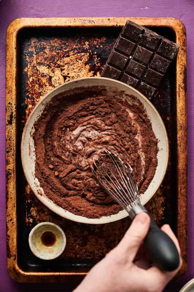 A hand uses a whisk to mix chocolate batter in a bowl—perfect for making chocolate ice cream. Nearby, a chocolate bar and a small cup of liquid rest on a worn baking sheet.