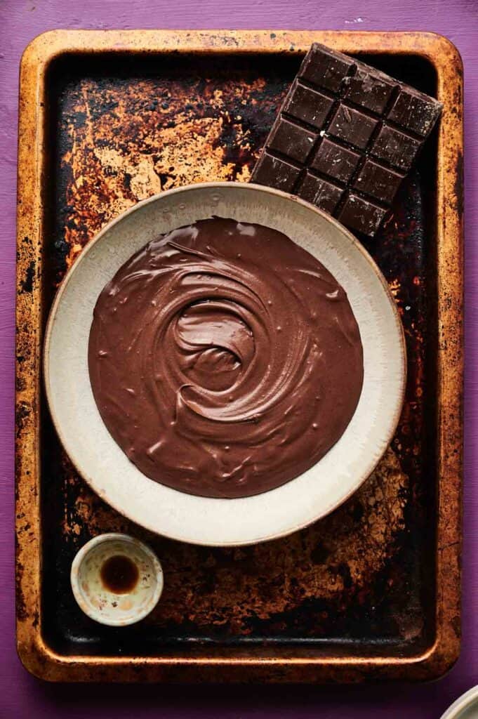 A bowl of melted chocolate sits on a baking tray next to a partially used chocolate bar and a small dish with liquid, perfect for making rich chocolate ice cream, viewed from above.