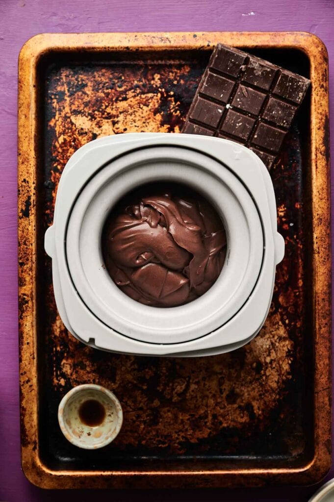 A container of chocolate ice cream mixture sits on a worn baking tray next to a bar of chocolate and a small cup of liquid on a purple surface.