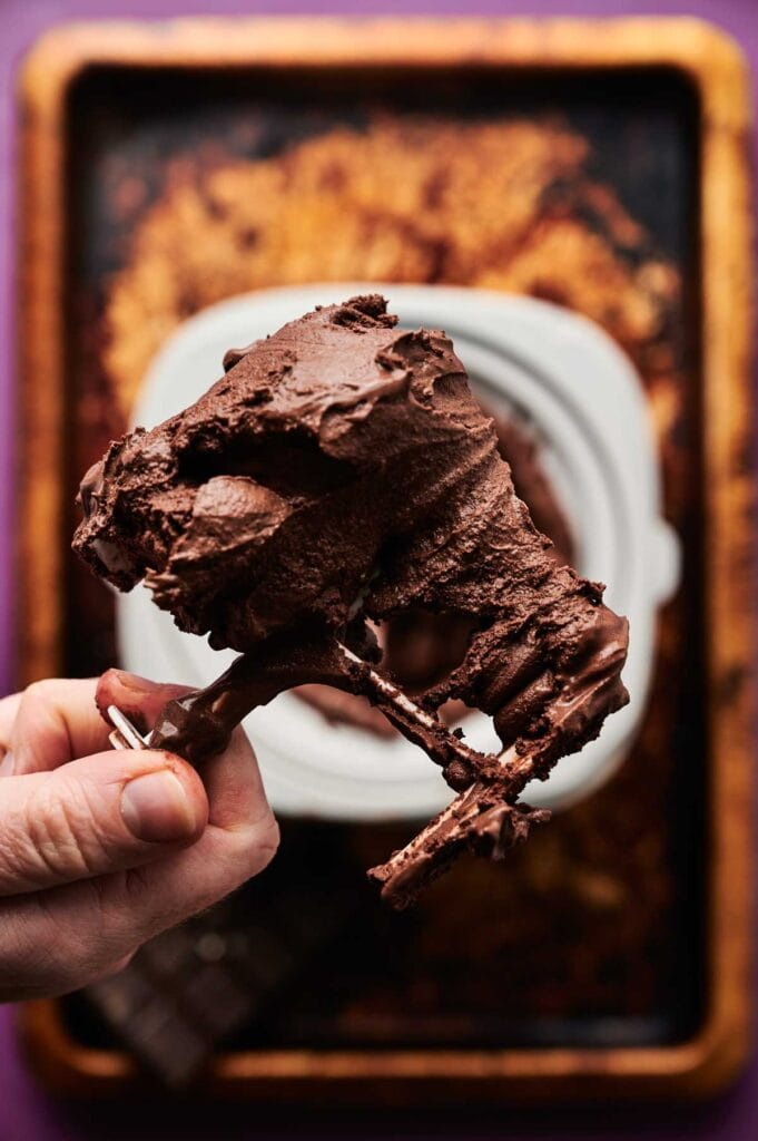 A hand holds a stand mixer whisk attachment covered in rich chocolate ice cream frosting above a mixing bowl and baking tray.