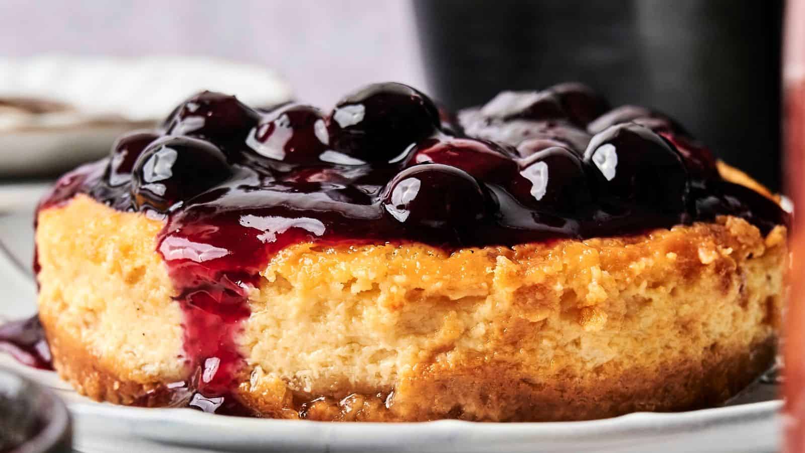 A round cheesecake topped with glossy blueberry sauce sits on a white plate.