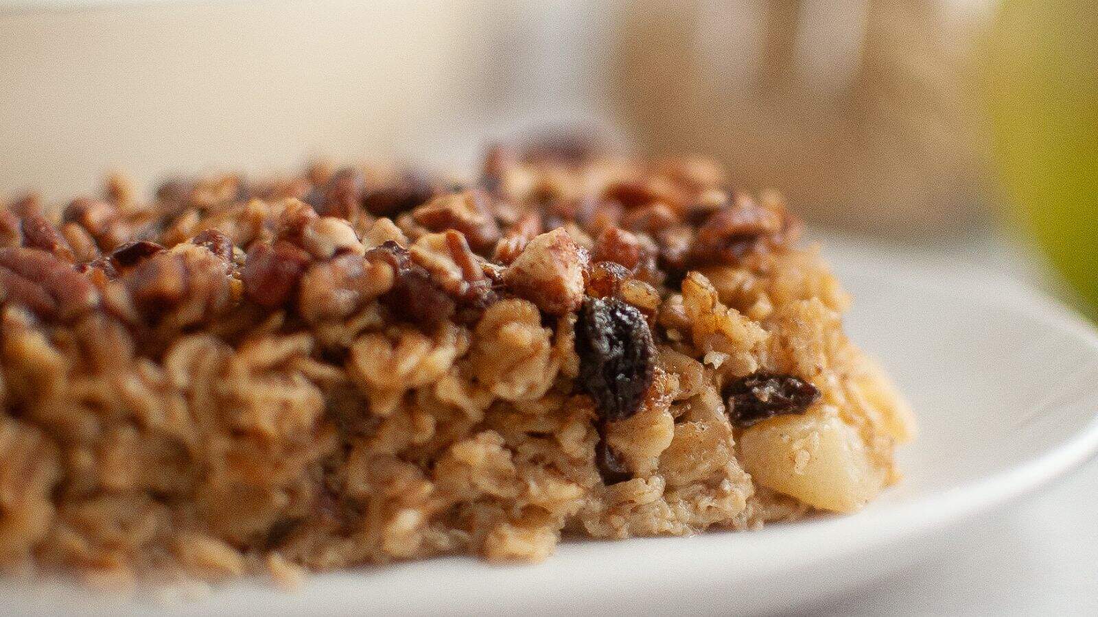 A close-up of a baked oatmeal bar with visible oats, nuts, and raisins on a white plate.