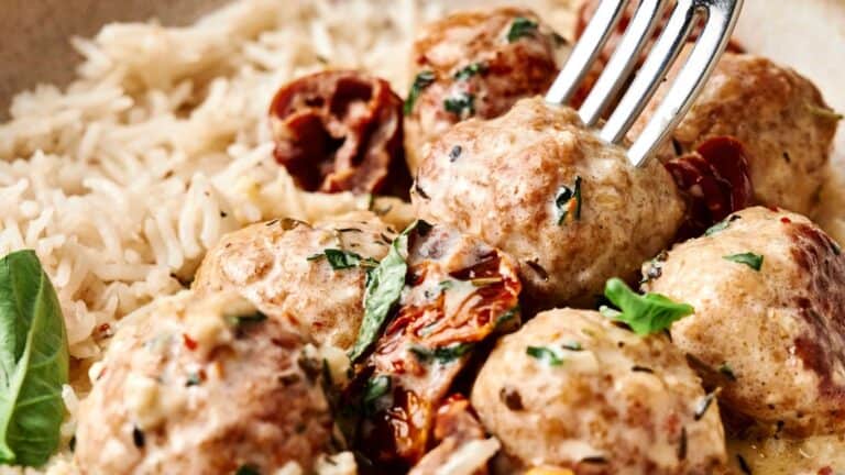 A close-up of marry me chicken meatballs in a creamy sauce with herbs and sun-dried tomatoes, served next to a portion of white rice.