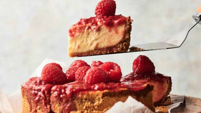 A slice of raspberry-topped cheesecake is being lifted from the whole cake, which has a graham cracker crust and fresh raspberries on top.