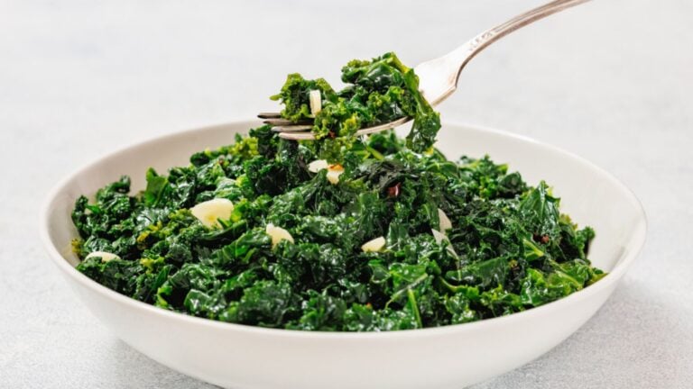 A white bowl filled with sautéed kale and garlic, with a fork lifting a portion from the bowl.