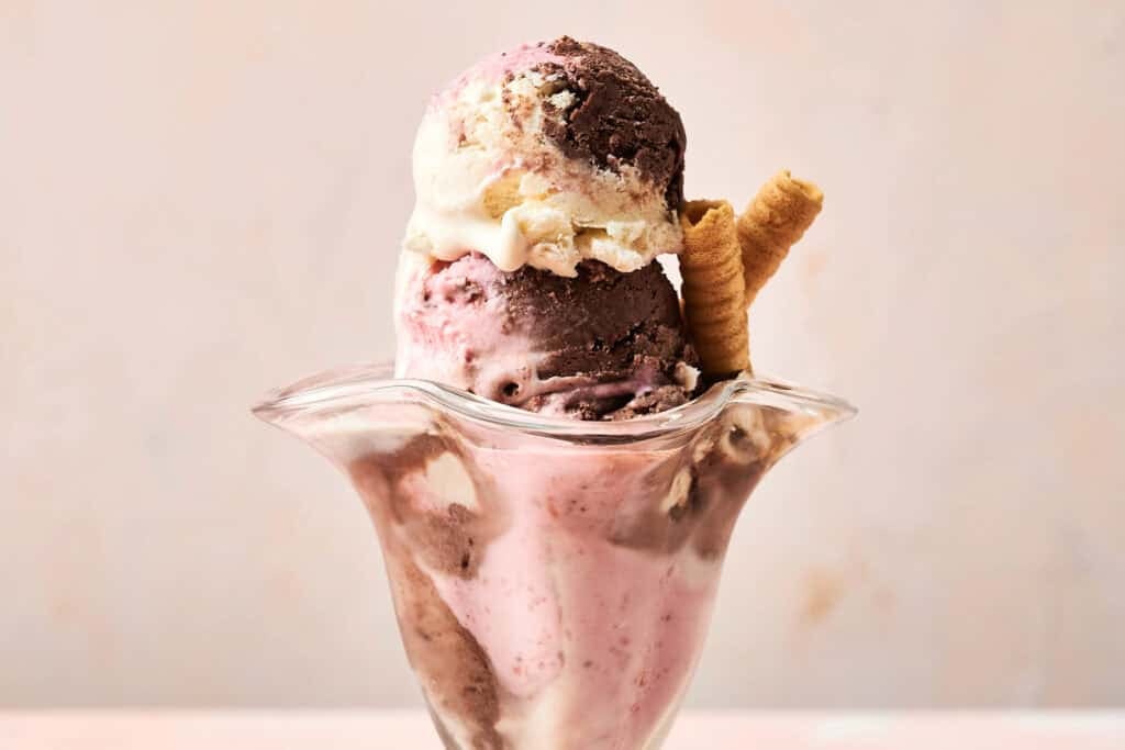 A glass sundae dish overflowing with scoops of homemade Neapolitan ice cream in chocolate, vanilla, and strawberry, garnished with a wafer cookie roll